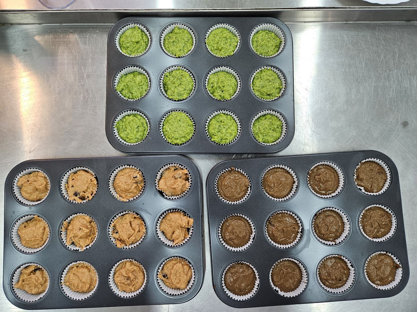 Trzy foremki na muffinki wypełnione surowym ciastem, zielonym, pomarańczowym i brązowym.