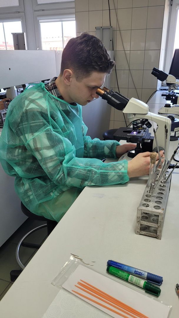 Uczeń w zielonym fartuchu laboratoryjnym oglądający preparat pod mikroskopem.