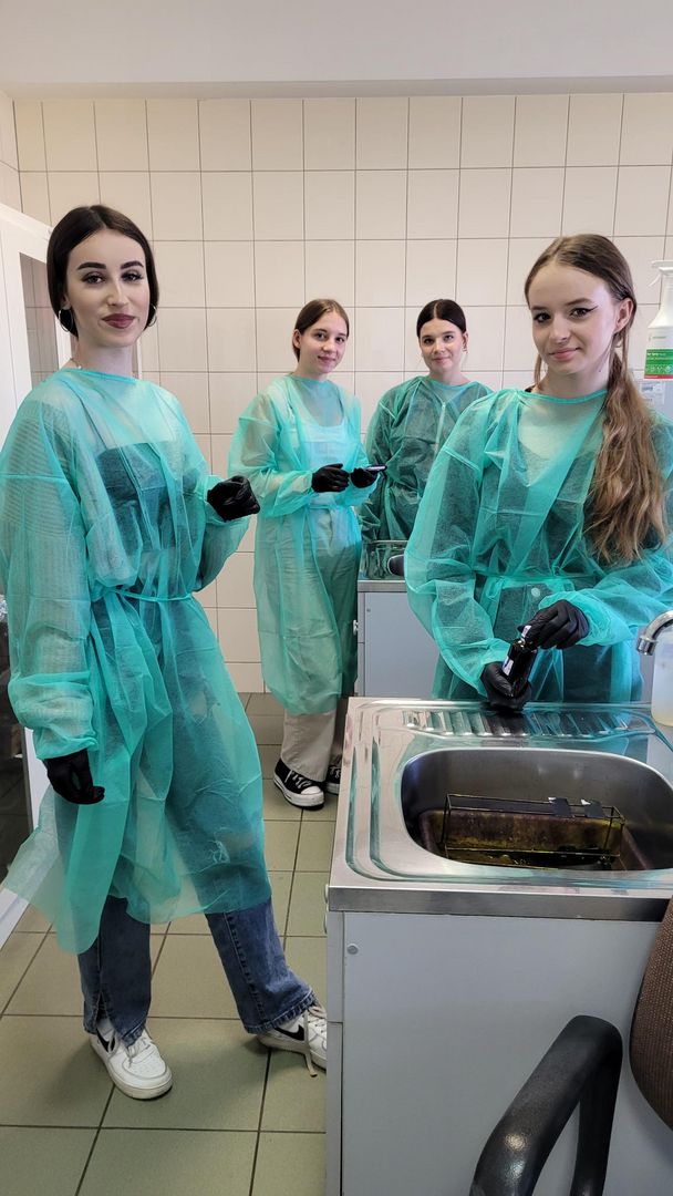 Cztery uczennice w zielonych fartuchach stojące przy zlewie i wykonujące barwienie preparatów mikroskopowych.